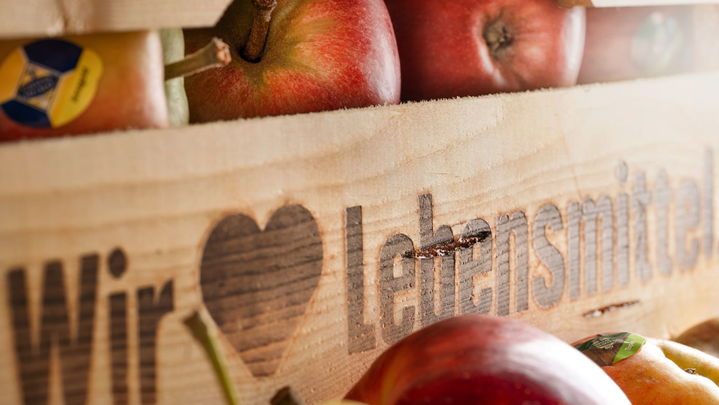 Bildausschnitt einer Holzkiste mit dem Aufschrift Wir ♥ Lebensmittel. Darin sind rote Äpfel mit Aufklebern 